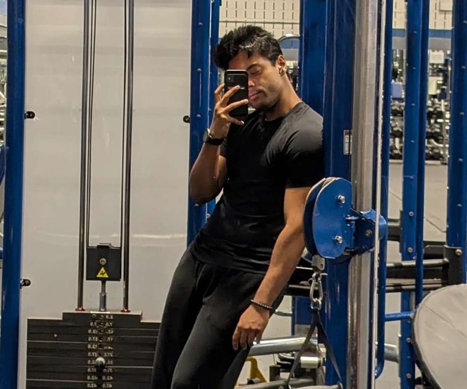 Aditya in the gym mirror, phone up, leaning on a blue cable machine.
